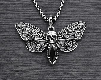 Op de afbeelding: Een zilverkleurige metalen ketting met een hanger in de vorm van een nachtvlinder, voorzien van een schedel als kop en een zwarte edelsteen in marquise-slijpvorm als lichaam. De vleugels zijn gegraveerd met hemelse patronen zoals sterren en manen.