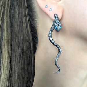Peut inclure: Une boucle d'oreille en forme de serpent argentée avec des yeux en pierres précieuses bleues. Le serpent est enroulé et a une surface texturée.