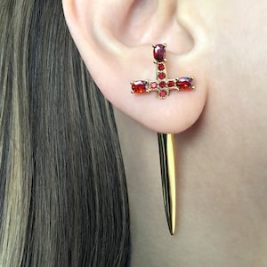 May include: Gold earring with a red crystal dagger design. The dagger is made of gold and has a red crystal cross at the top. The earring is a statement piece and would be perfect for a gothic or punk style.