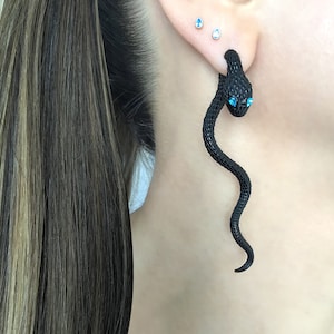 Könnte beinhalten: Schwarzer schlangenförmiger Ohrring mit blauen Edelsteinaugen. Der Ohrring wird am Ohr einer Person getragen.