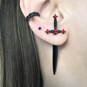 May include: Black sword-shaped earrings with red crystal accents. The earrings are worn in the earlobe and helix piercing.