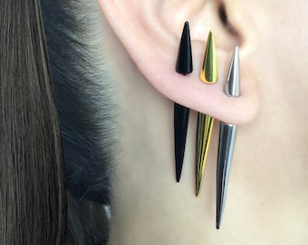 May include: Three different colored spike earrings. The earrings are black, gold, and silver. The earrings are all the same shape, but different sizes. The earrings are worn in a person's ear.