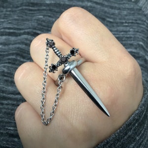 Könnte beinhalten: Ein silberner Ring mit einem Schwertmuster und schwarzen Edelsteinen. Das Schwert hat eine Kette daran.
