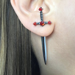 May include: A silver dagger earring with red gemstone accents. The earring is designed to look like a sword with a pointed tip.