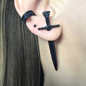 May include: Black sword-shaped earrings with intricate details. The earrings are made of metal and have a Celtic knot design. The earrings are worn in the earlobe and helix piercing.
