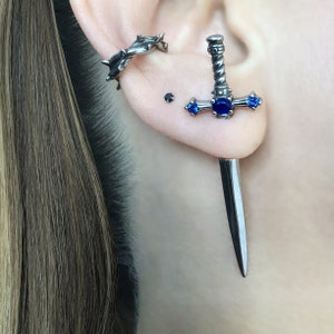 May include: A pair of silver earrings, one is a sword with blue gemstones and the other is a thorned hoop.