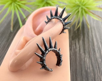 May include: A pair of silver ear cuffs with a spiky, crown-like design. The cuffs are shown on a model ear.