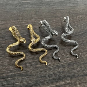 Peut inclure: Trois paires de boucles d'oreilles en forme de serpent dans des finitions or, argent et noir. Les boucles d'oreilles sont en métal et ont une surface texturée.
