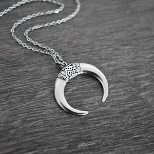 May include: Silver-toned crescent moon pendant necklace. The pendant features a detailed, patterned band at the top and curved, polished horns. The necklace chain is delicate and complements the pendant's design. A stylish piece of jewelry.
