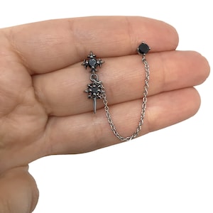 May include: A silver-toned ear cuff with a black gemstone stud and a chain connecting to a decorative earring. The decorative earring features a cross-like design with a central black gemstone and a small, pointed element.