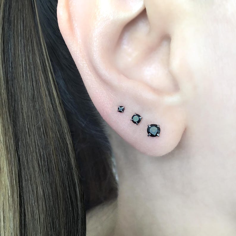 May include: Three black gemstone earrings of different sizes, set in silver, worn in the earlobe.
