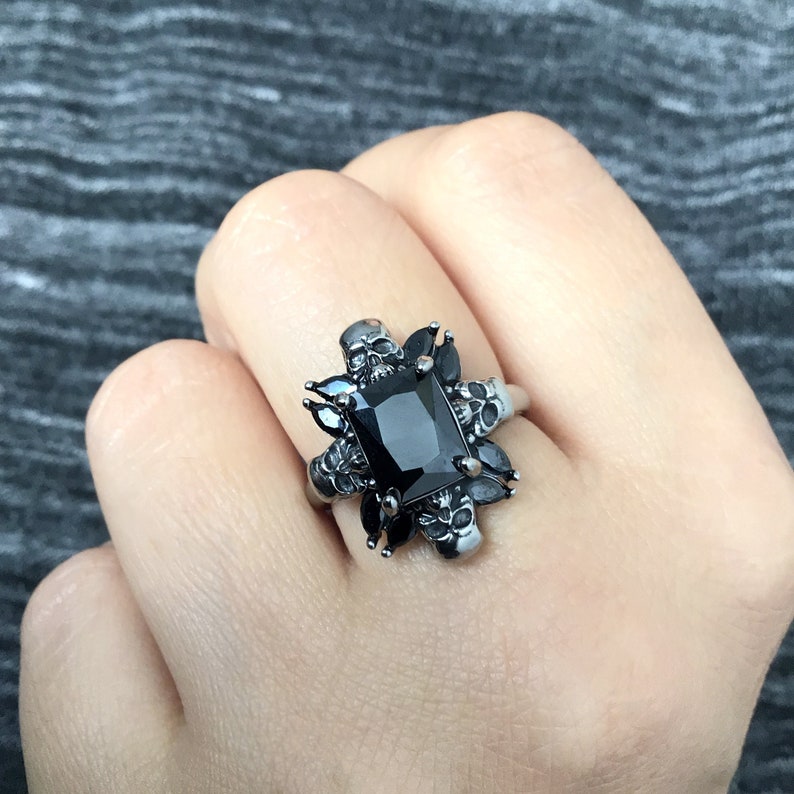 May include: A black onyx ring with a square-shaped stone surrounded by silver skulls and black accents.