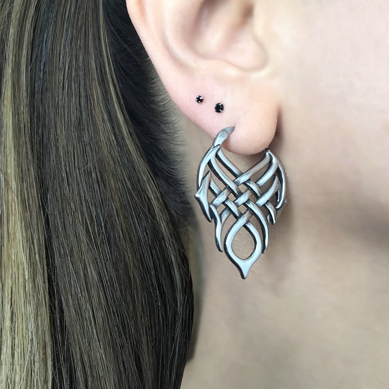 May include: Silver hoop earrings with a detailed, intricate, woven design. The earrings are shaped like a teardrop and have a dangling, open-ended design.