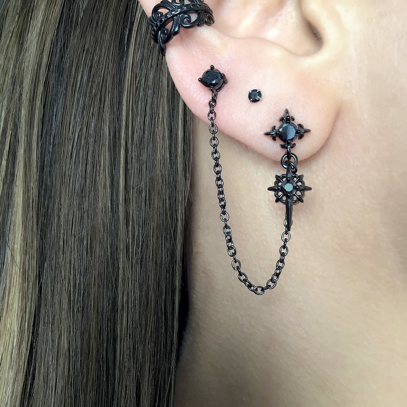 May include: Black ear cuff and earrings set. The ear cuff features an ornate design. The earrings include a stud, a small stud, and a chain that connects to a starburst-shaped earring with a black gem.