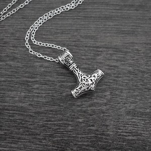 May include: A silver-colored pendant necklace featuring a detailed Thor's hammer design. The pendant is intricately carved with ornamental patterns and hangs from a delicate chain. The necklace is displayed on a dark wooden surface.