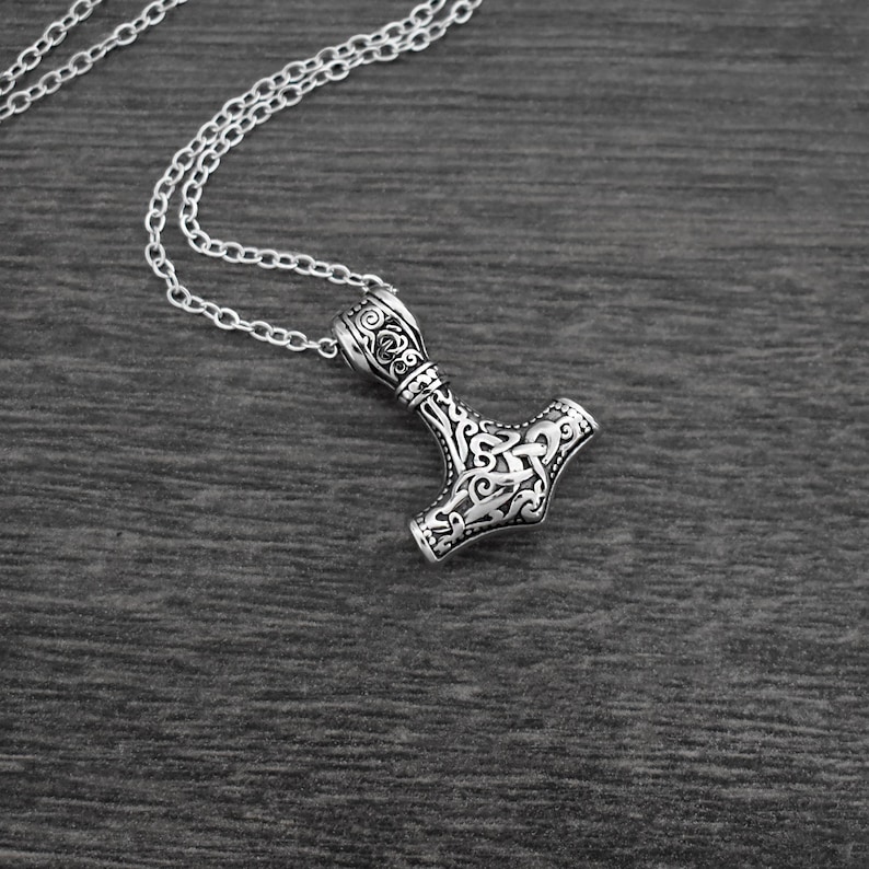 May include: A silver-colored pendant necklace featuring a detailed Thor's hammer design. The pendant is intricately carved with ornamental patterns and hangs from a delicate chain. The necklace is displayed on a dark wooden surface.