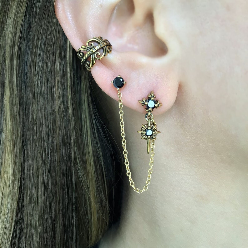 May include: An ear cuff and two earrings are shown. The ear cuff is a bronze color with an ornate design. One earring is a black stud with a gold chain that connects to a second earring with a starburst design and a black gem.
