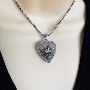 May include: A silver-toned heart-shaped locket pendant suspended from a matching box chain. The locket features an engraved North Star design surrounded by small stars and a beaded border, finished with an oxidised effect for depth and detail.
