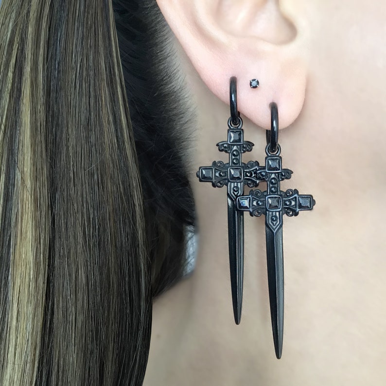 May include: A pair of black dangle earrings featuring a sword-shaped cross design with black cubic zirconia stones.