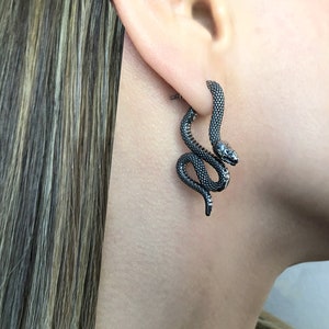 May include: A pair of silver snake earrings. The earrings are shaped like a snake with its head curled up. The snake is detailed with scales and has a black finish.