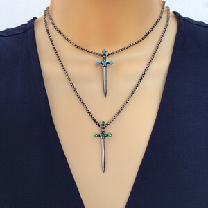 May include: A silver chain necklace with two sword pendants. The top pendant has turquoise stones and the bottom pendant has green stones.