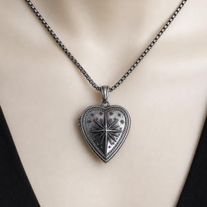 May include: A dark silver-toned heart-shaped locket pendant suspended from a matching box chain. The locket features an engraved North Star design surrounded by smaller star motifs and a beaded border, showcasing a weathered, oxidised metal finish.