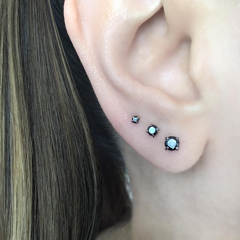 May include: Three black diamond stud earrings of different sizes. The earrings are set in silver and are worn in the earlobe.