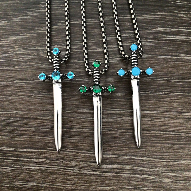 May include: Three silver sword necklaces with different coloured gemstones. The swords have blue and green gemstones on the hilt. The necklaces are on a wooden surface.