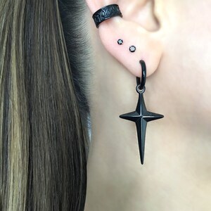 May include: Black star-shaped earring with a hoop and a black cross pendant. The earring is worn in the lower lobe of the ear. There are two small black studs in the upper lobe of the ear. The earring is worn by a person with long brown hair.