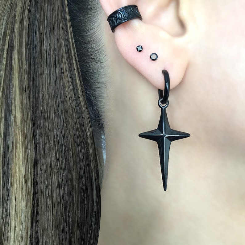 May include: Black star-shaped earring with a hoop and a black cross pendant. The earring is worn in the lower lobe of the ear. There are two small black studs in the upper lobe of the ear. The earring is worn by a person with long brown hair.