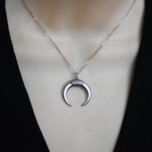 May include: Silver-toned necklace featuring a crescent-shaped pendant. The pendant has a detailed top section and a smooth, curved horn design. The necklace is displayed on a mannequin, with a black garment in the background.