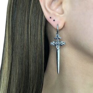 May include: Silver sword cross dangle earring with black cubic zirconia stones. The earring is hanging from a hoop.