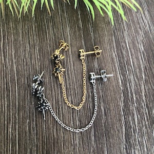 May include: Three pairs of earrings in gold, silver, and black. Each pair features a stud and a chain that connects to a second stud. The gold and black earrings have a starburst design with a black gem. The silver earrings have a floral design.