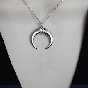 May include: A silver-toned necklace featuring a crescent-shaped pendant. The pendant has a textured design at the top and a smooth, curved horn shape. The chain is a delicate link style, suitable for everyday wear.