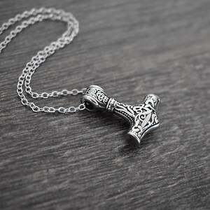 May include: A silver-colored pendant necklace featuring a detailed hammer design, likely inspired by Norse mythology. The hammer pendant is intricately decorated with swirling patterns and is suspended from a delicate chain. The necklace rests on a dark wooden surface.