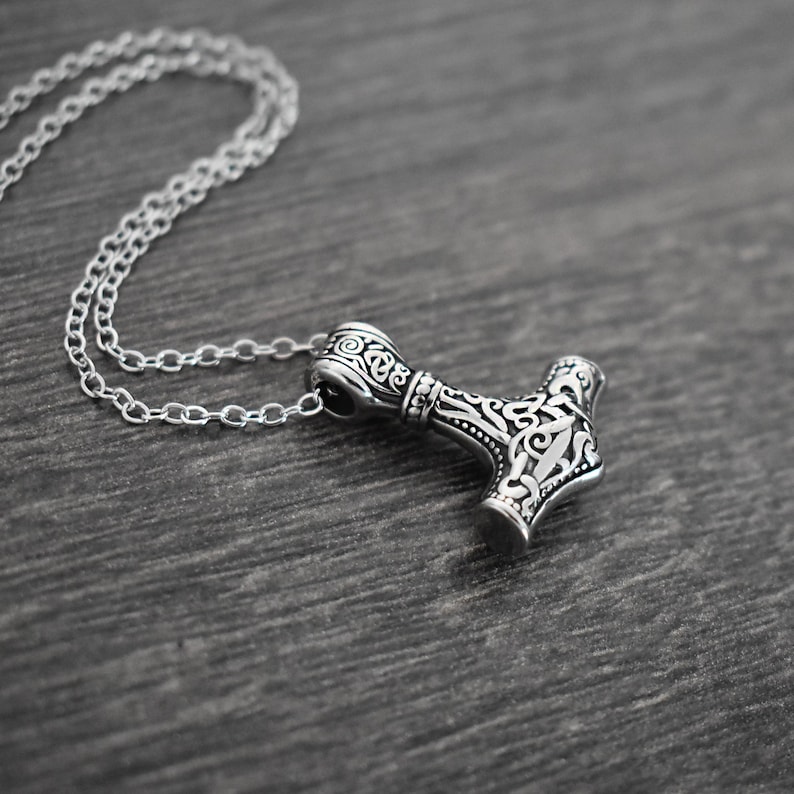 May include: A silver-colored pendant necklace featuring a detailed hammer design, likely inspired by Norse mythology. The hammer pendant is intricately decorated with swirling patterns and is suspended from a delicate chain. The necklace rests on a dark wooden surface.