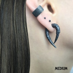 Peut inclure: Une paire de boucles d'oreilles noires textur&eacute;es en forme de griffe. Les boucles d'oreilles ont la forme d'une griffe incurv&eacute;e avec une pointe pointue. Les boucles d'oreilles sont port&eacute;es au lobe de l'oreille et au piercing de l'h&eacute;lix.