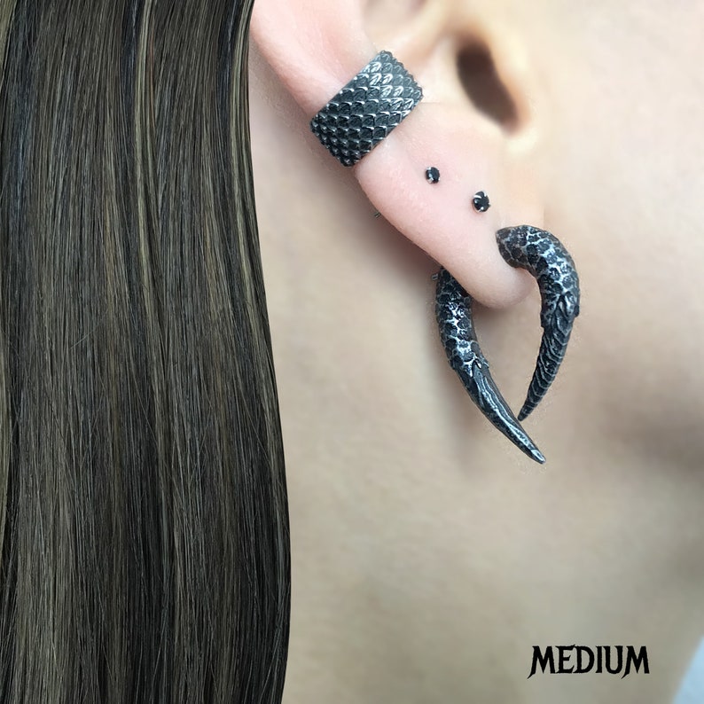 Peut inclure: Une paire de boucles d'oreilles noires textur&eacute;es en forme de griffe. Les boucles d'oreilles ont la forme d'une griffe incurv&eacute;e avec une pointe pointue. Les boucles d'oreilles sont port&eacute;es au lobe de l'oreille et au piercing de l'h&eacute;lix.