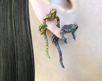 Peut inclure: Une paire de boucles d'oreilles avec un motif de lézard doré et noir. Les lézards sont positionnés comme s'ils grimpaient sur le lobe de l'oreille.