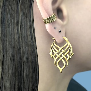 May include: Gold hoop earrings with a Celtic knot design. The earrings are large and dangle from the earlobe. The earrings are worn by a person with long brown hair.