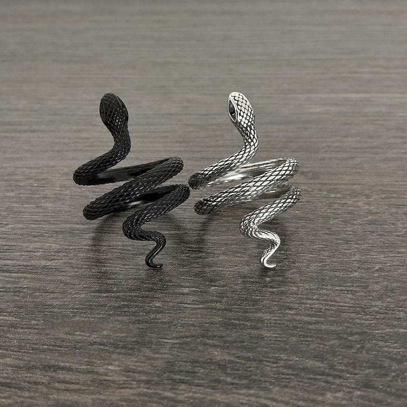Puede incluir: Dos anillos de serpiente, uno negro y otro plateado, est&aacute;n enroscados y mirando hacia arriba. Los anillos est&aacute;n sobre una superficie de madera oscura.