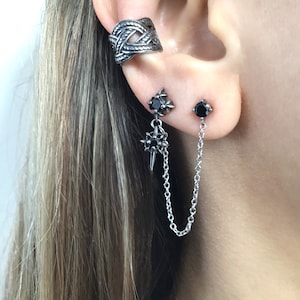 May include: A silver-toned ear cuff and two earrings are displayed on an ear. The ear cuff has a woven design. One earring is a stud with a black gem, and the other has a starburst design with a chain that connects to a second black gem stud.