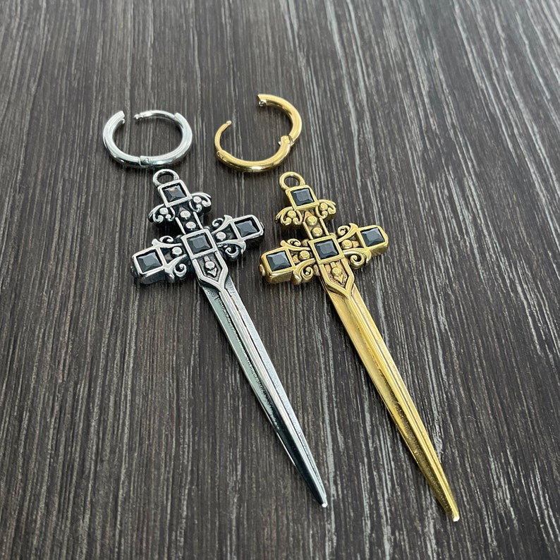 May include: Two silver and gold sword-shaped cross earrings with black gemstones. The earrings are on a wooden surface.