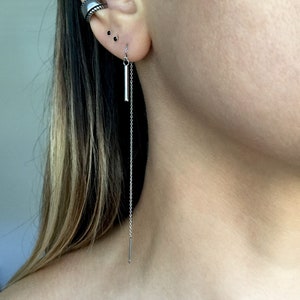 May include: A silver chain earring with a small silver bar hanging from it. The earring is worn in the lower lobe of the ear.