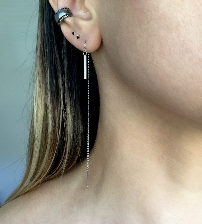 May include: A silver chain earring with a small silver bar hanging from it. The earring is worn in the lower lobe of the ear.
