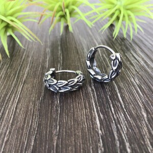 Puede incluir: Un par de pendientes de aro de plata con un dise&ntilde;o de hojas. Los pendientes est&aacute;n sobre una superficie de madera.