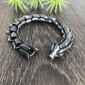 Puede incluir: Un brazalete de drag&oacute;n plateado con un dise&ntilde;o texturizado y escamoso. El brazalete est&aacute; abierto y tiene un cierre en el lateral.