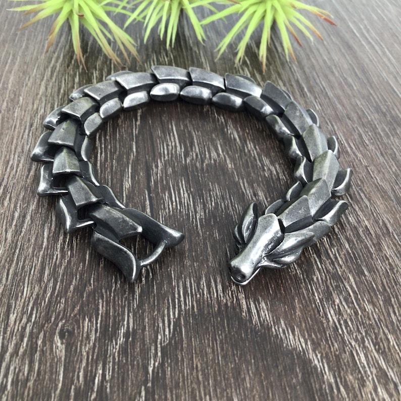 Puede incluir: Un brazalete de drag&oacute;n plateado con un dise&ntilde;o texturizado y escamoso. El brazalete est&aacute; abierto y tiene un cierre en el lateral.