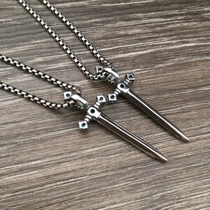 May include: Two silver sword pendants with blue gemstone accents on silver chains. The swords are detailed with intricate designs and have a pointed tip.