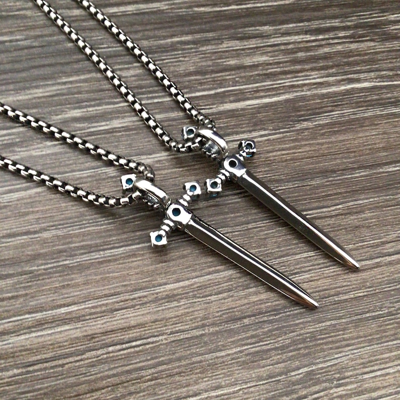 May include: Two silver sword pendants with blue gemstone accents on silver chains. The swords are detailed with intricate designs and have a pointed tip.